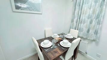 A dining area with a rectangular table set for four, white chairs, a framed picture on the wall, and a window with patterned curtains.