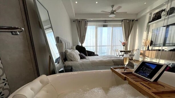 A modern hotel room with a large bathtub, a bed with a headboard, a desk with a computer, and a large window with curtains. - Luxury Seaview Studio Apartment - RAK Al Marjan Island (Ras Al-Khaimah)