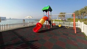 A playground with a slide and swings, a white picket fence, and a view of the sea. - Luxury Seaview Studio Apartment - RAK Al Marjan Island (Ras Al-Khaimah)