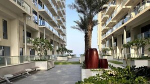 A modern multi-story residential building with balconies, a central palm tree, and a paved courtyard with benches and planters. - Luxury Seaview Studio Apartment - RAK Al Marjan Island (Ras Al-Khaimah)