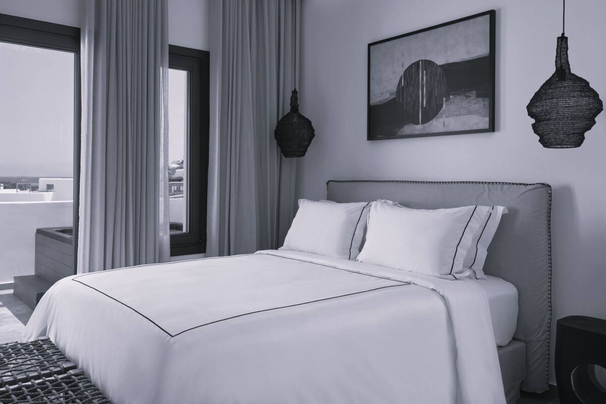 A modern hotel room with a large bed, a view through the window, and a painting on the wall.