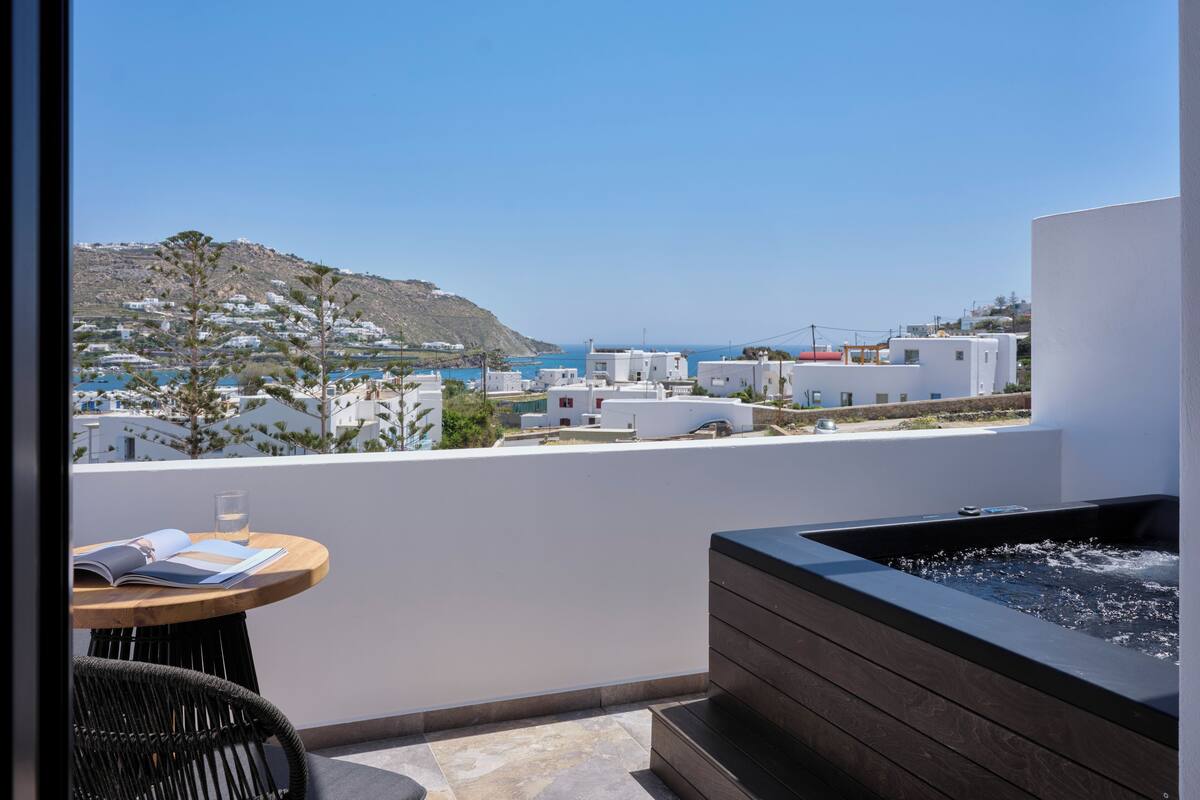 A balcony with a view of a coastal town, a hot tub, and a table with a book and glass.