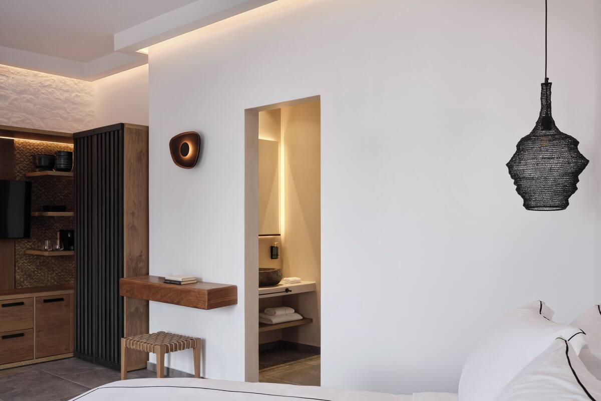 A modern bedroom with a bed, a wooden bench, a small stool, a bedside table with a book, a mirror, and a hanging light fixture.