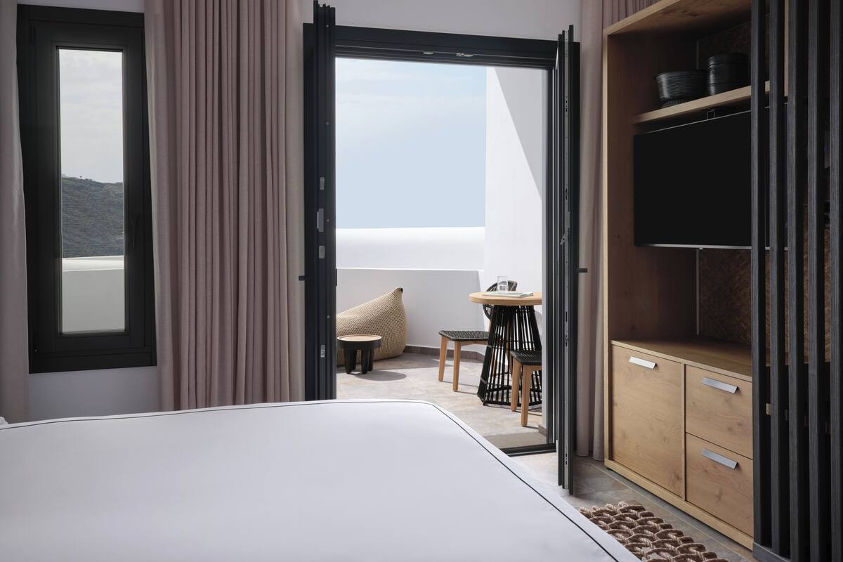 A modern hotel room with a large bed, a balcony with a view, a small table with a chair, and a TV on a wooden cabinet.