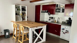 Pieni, käytännöllinen keittiö, jossa on puinen pöytä, kaksi tuolia, valkoiset lasiovet, sekä punainen kaappi, jonka sisällä on mikroaaltouuni ja kahvinkeitin.