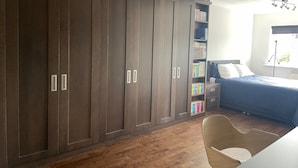 Une chambre avec une grande armoire, une table de chevet avec des livres, un lit recouvert d’une couverture bleue et une chaise.