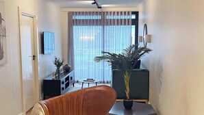 A modern living room with a sofa, a dining table, a potted plant, and a TV mounted on the wall.