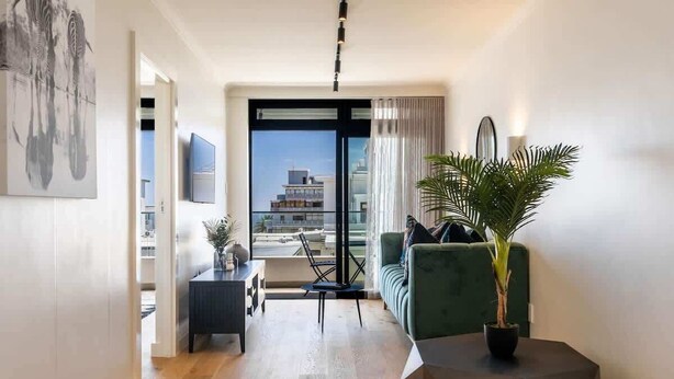 A modern living room with a sofa, a coffee table, and a potted plant. There is a view of buildings through a glass door.