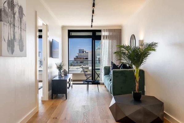 A modern living room with a sofa, a coffee table, and a potted plant. There is a view of buildings through a glass door.