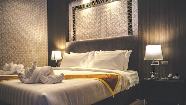 A hotel room with a neatly made bed, a bedside lamp, a telephone, and a wall with a patterned wallpaper.