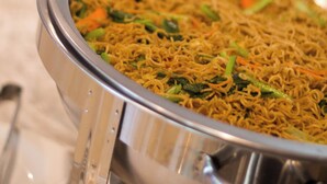 A buffet with multiple metal serving dishes containing a noodle dish with vegetables.