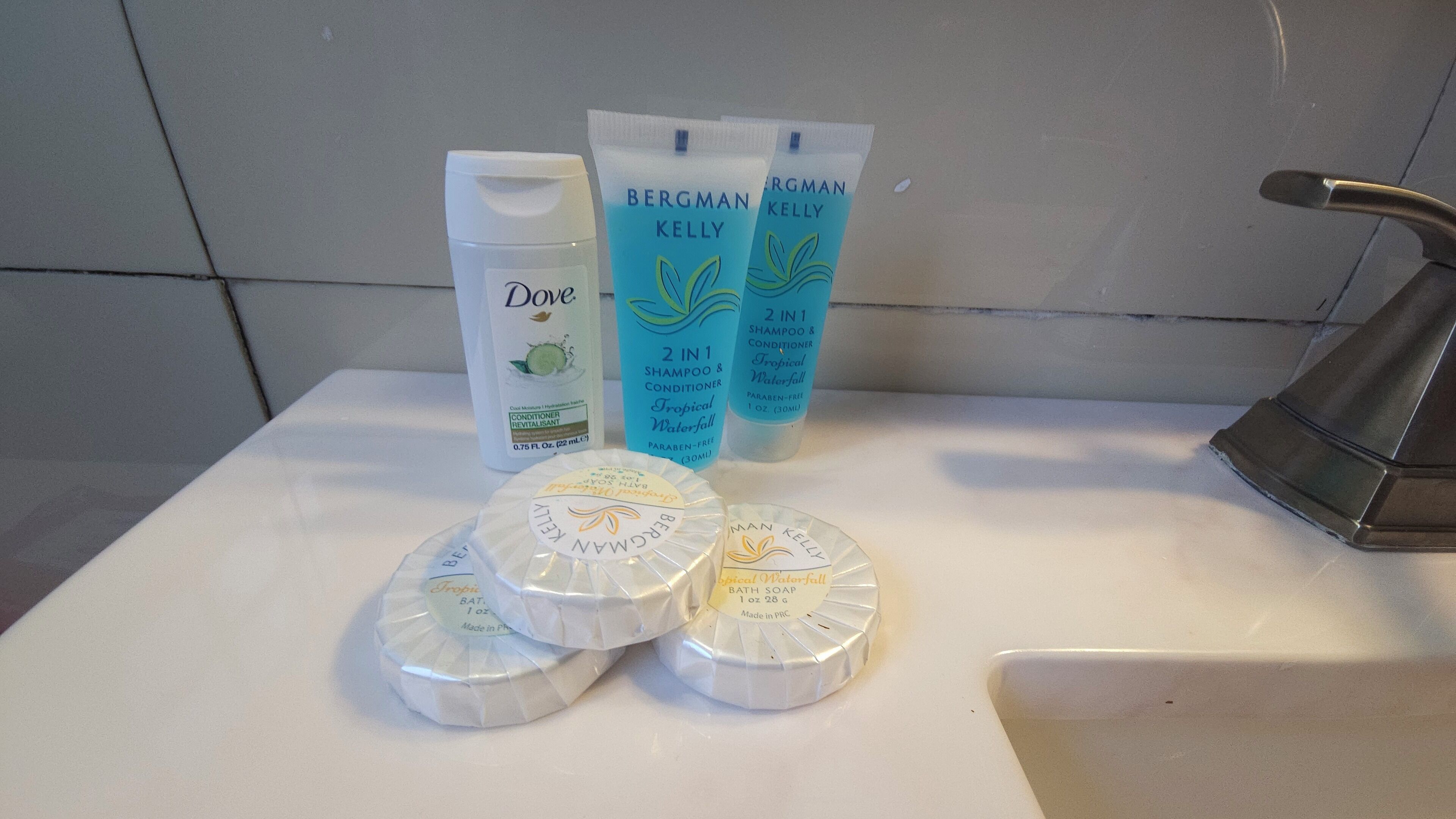 Et badeværelsesvask med toiletartikler, heriblandt en flaske Dove conditioner, to tuber Bergman Kelly shampoo og conditioner, samt tre Bergman Kelly sæber.