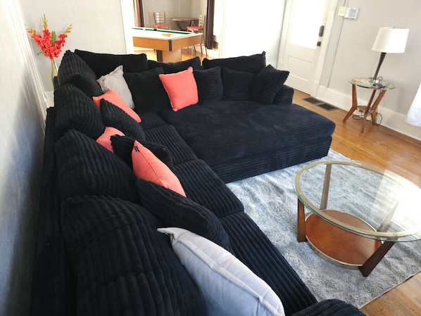 A sectional sofa with multiple cushions, a glass coffee table, and a dining area in the background. - Charming 4-Bedroom Historic Home w/ Game Room + Backyard BBQ (Goldsboro)
