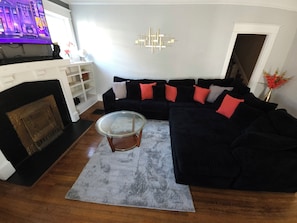 A living room with a black sectional sofa, a glass coffee table, a fireplace, and a modern wall art piece. - Charming 4-Bedroom Historic Home w/ Game Room + Backyard BBQ (Goldsboro)