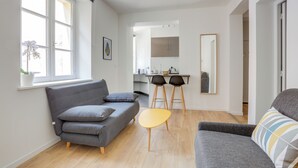 A modern living room with a grey sofa, a yellow table, and a grey armchair. There is a kitchen area in the background with a dining table and chairs. - Le Recollet - Versailles (Versailles)