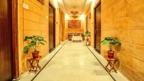 A hotel hallway with wooden walls, potted plants, a reception desk, and a bed in the distance. - Hotel Mantra Heritage Jaisalmer (Jaisalmer)