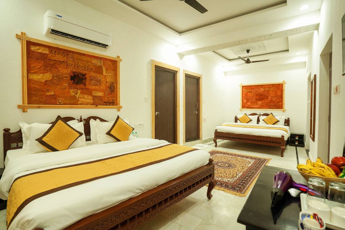 A hotel room with two beds, a large decorative wall hanging, an air conditioning unit, and a small table with fruits and a hairdryer.