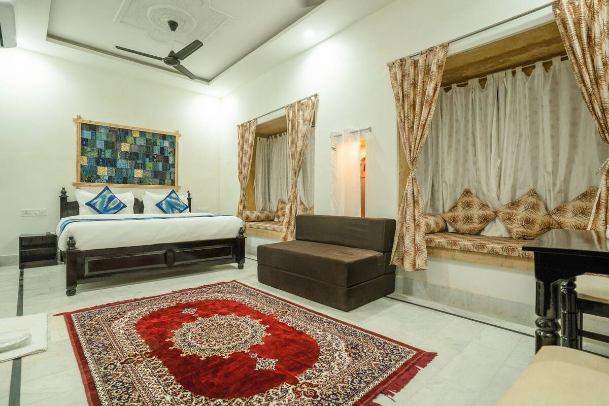 A bedroom with a bed, a sofa, a red patterned rug, a ceiling fan, and curtains.