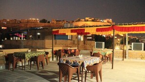An outdoor dining area with tables and chairs, set against a backdrop of a historic city wall and buildings. - Hotel Mantra Heritage Jaisalmer (Jaisalmer)