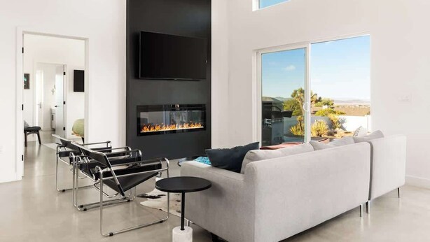 A modern living room with a fireplace, a sofa, a coffee table, and a large window offering a view of the outdoors. - No Vacancy • New Home, Game Room, Spa, Hammocks (Joshua Tree)
