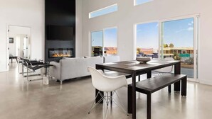 A spacious living area with a modern fireplace, a dining table, and large windows offering a view of the landscape. - No Vacancy • New Home, Game Room, Spa, Hammocks (Joshua Tree)