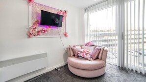 Una sala de estar moderna con un sofá rosa, un televisor de pantalla plana montado en una pared decorativa y ventanas amplias que ofrecen vistas de la ciudad.