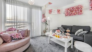 Una sala de estar moderna con un sofá gris, una mesa de centro blanca con pastel y aperitivos, y un sofá rosa adornado con flores.