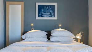 Una cama bien hecha con ropa blanca, dos lámparas de lectura fijadas a la pared y una obra de arte con temática náutica enmarcada sobre la cama.