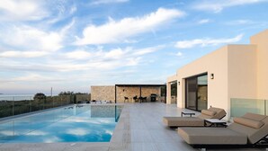 A modern outdoor pool area with a stone wall, lounge chairs, and a barbecue setup. - Villa Kafkales | Heated Pool (Chania)