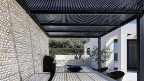 An outdoor patio with a stone wall, a grill, a table with chairs, and a pergola overhead. - Villa Kafkales | Heated Pool (Chania)