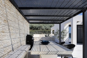 An outdoor patio with a stone wall, a grill, a table with chairs, and a pergola overhead.