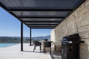 An outdoor patio with a grill, chairs, and a view of the sea and hills.