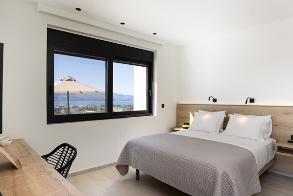 A modern bedroom with a large bed, a desk, and a window with a view of the sea.