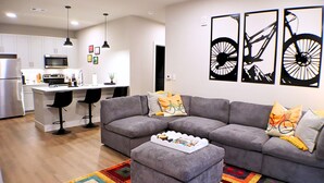 Una sala moderna con un sofá seccional gris, una mesa de centro y una alfombra colorida. Hay una cocina con armarios blancos y un microondas.