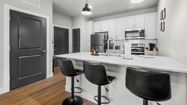 Una cocina moderna con alacenas blancas, una isla grande con taburetes y electrodomésticos como microondas y refrigerador.