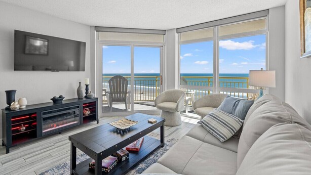 A spacious living room with a large window overlooking the ocean, a flat-screen TV mounted on the wall, and a comfortable seating area with a sofa and chairs. - Oceanfront King Bed 2BR 2BA Condo with WD Kitchen (North Myrtle Beach)