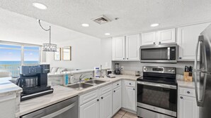 A modern kitchen with white cabinets, stainless steel appliances, a coffee maker, and a view of the ocean. - Oceanfront King Bed 2BR 2BA Condo with WD Kitchen (North Myrtle Beach)