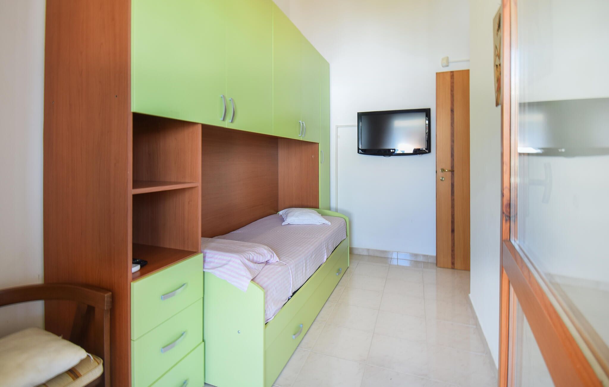 A small, single-bed room with a green wardrobe, a wooden chair, a television mounted on the wall, and a refrigerator.