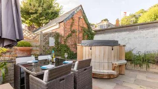 An outdoor patio with wicker furniture, a hot tub, and a brick building in the background. - Apartmentsleeps3-garden-hottub-parking (Padstow)