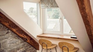 A small, cozy room with a sloped ceiling, a stone wall, and a window overlooking a garden. There are two wooden chairs and a small table. - Apartmentsleeps3-garden-hottub-parking (Padstow)