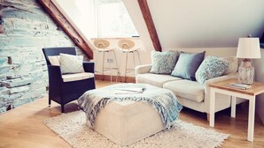 A loft-style living room with a high ceiling, exposed wooden beams, a white sofa, a dark wicker chair, a small ottoman, a white side table with a lamp, and a rustic brick wall. - Apartmentsleeps3-garden-hottub-parking (Padstow)