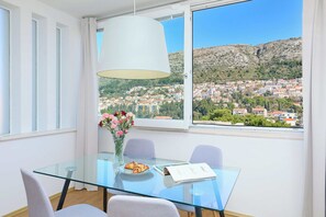 A dining area with a glass table, chairs, a vase with flowers, and a view of a town with houses and hills. - Cityview Apartment (Dubrovnik)