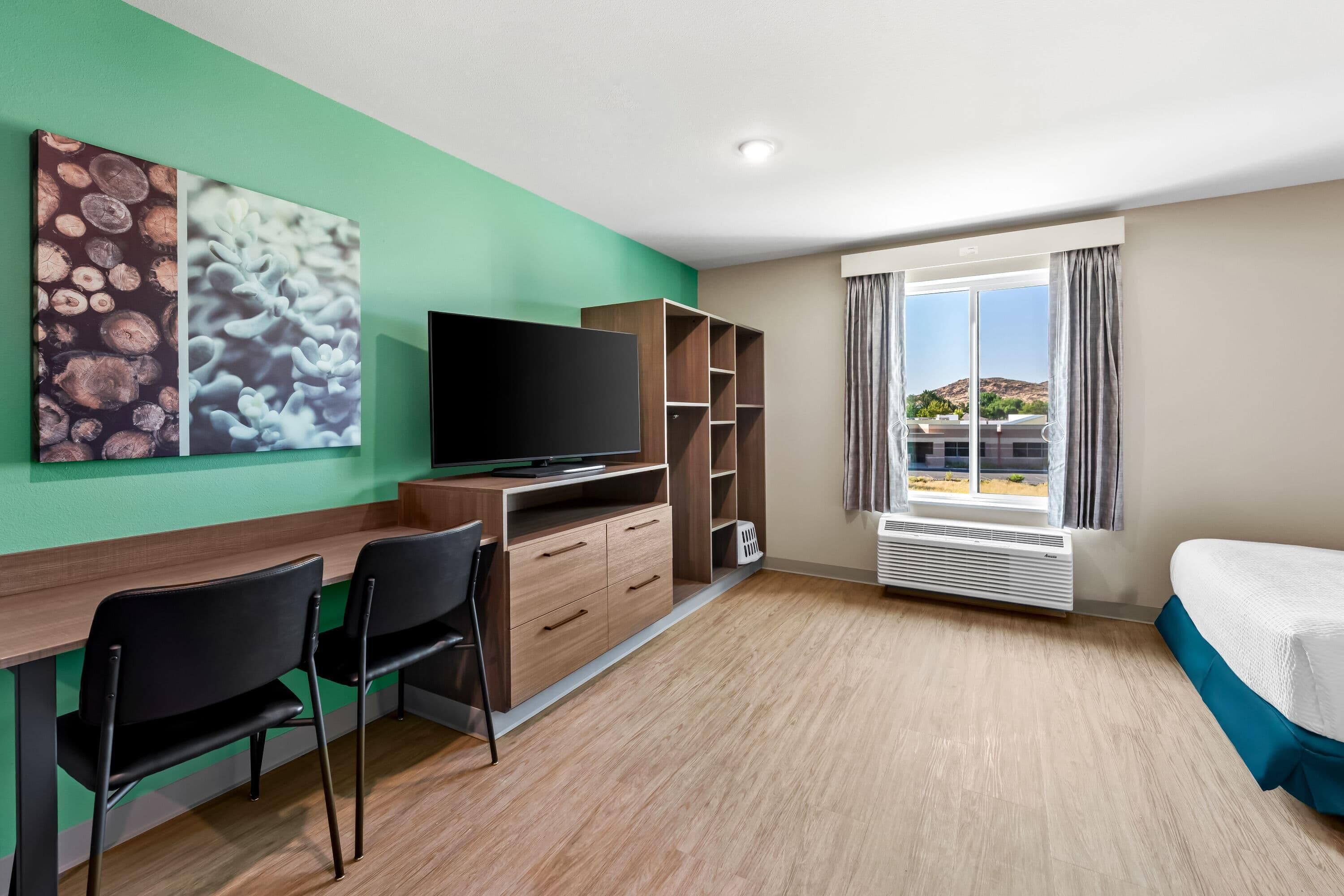 A hotel room with a wooden desk, two chairs, a flat-screen TV, a bed with a teal bedspread, a large window with curtains, and artwork on the wall.