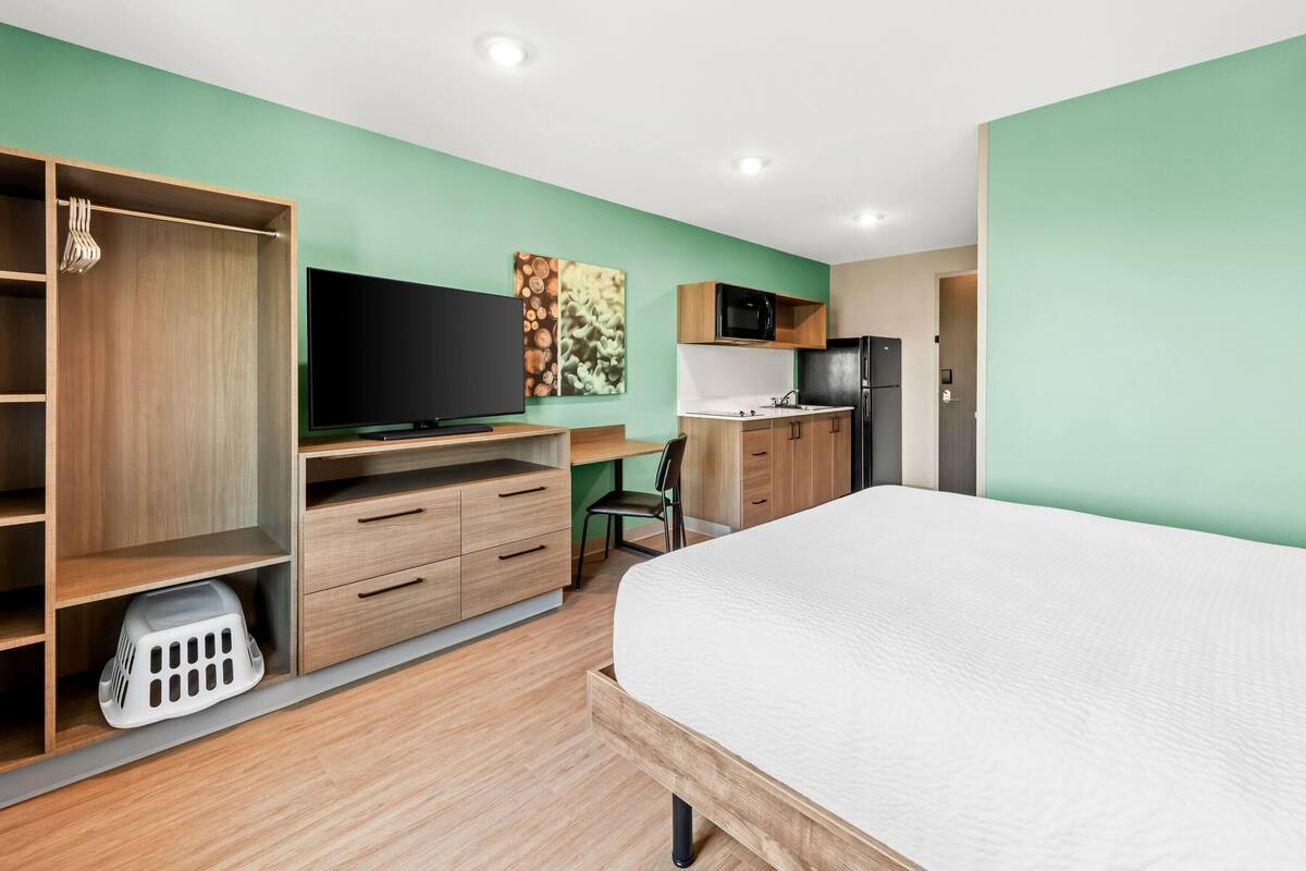 A modern room with a bed, wooden wardrobe, TV, kitchenette, and a painting on the wall.