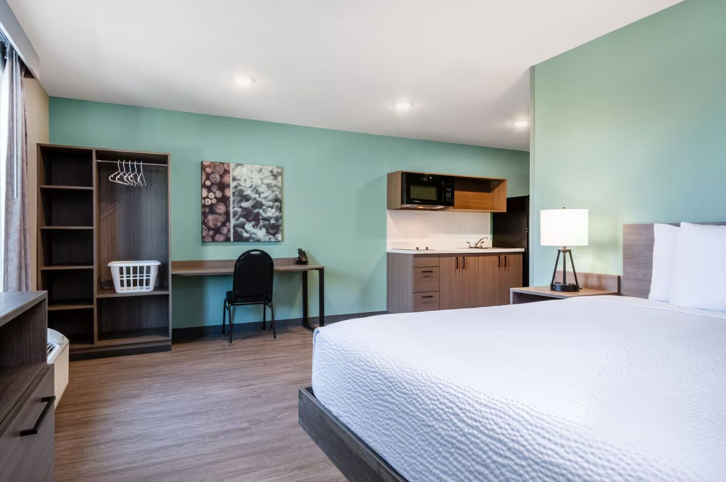 A hotel room with a bed, a desk, a chair, a wardrobe, a microwave, a sink, and a nightstand with a lamp.