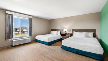 Habitación de hotel con dos camas, una ventana con cortinas, una mesita de noche con lámpara y vistas a una montaña y edificios.
