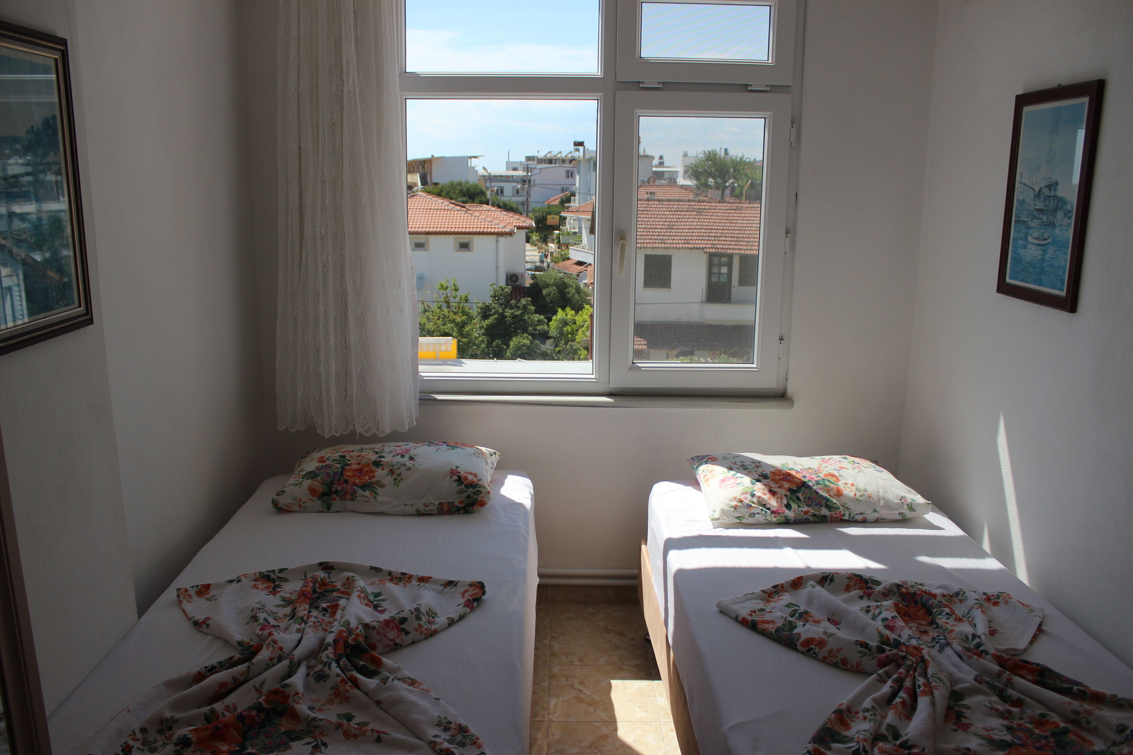 A room with two beds, a window with a view of buildings, and a framed picture on the wall.