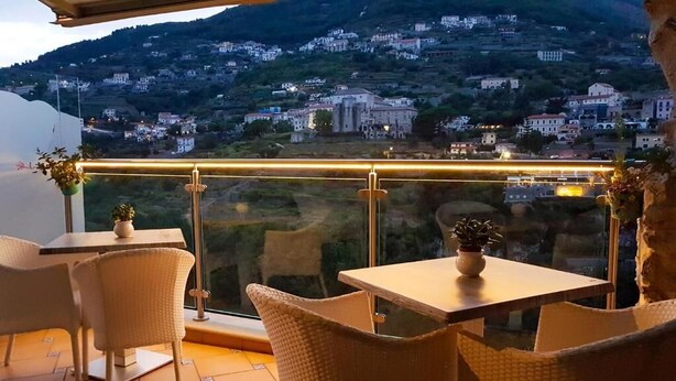 A terrace with wicker furniture and potted plants, overlooking a hillside town with a bridge in the distance. - Le perle d'italia (Ravello)