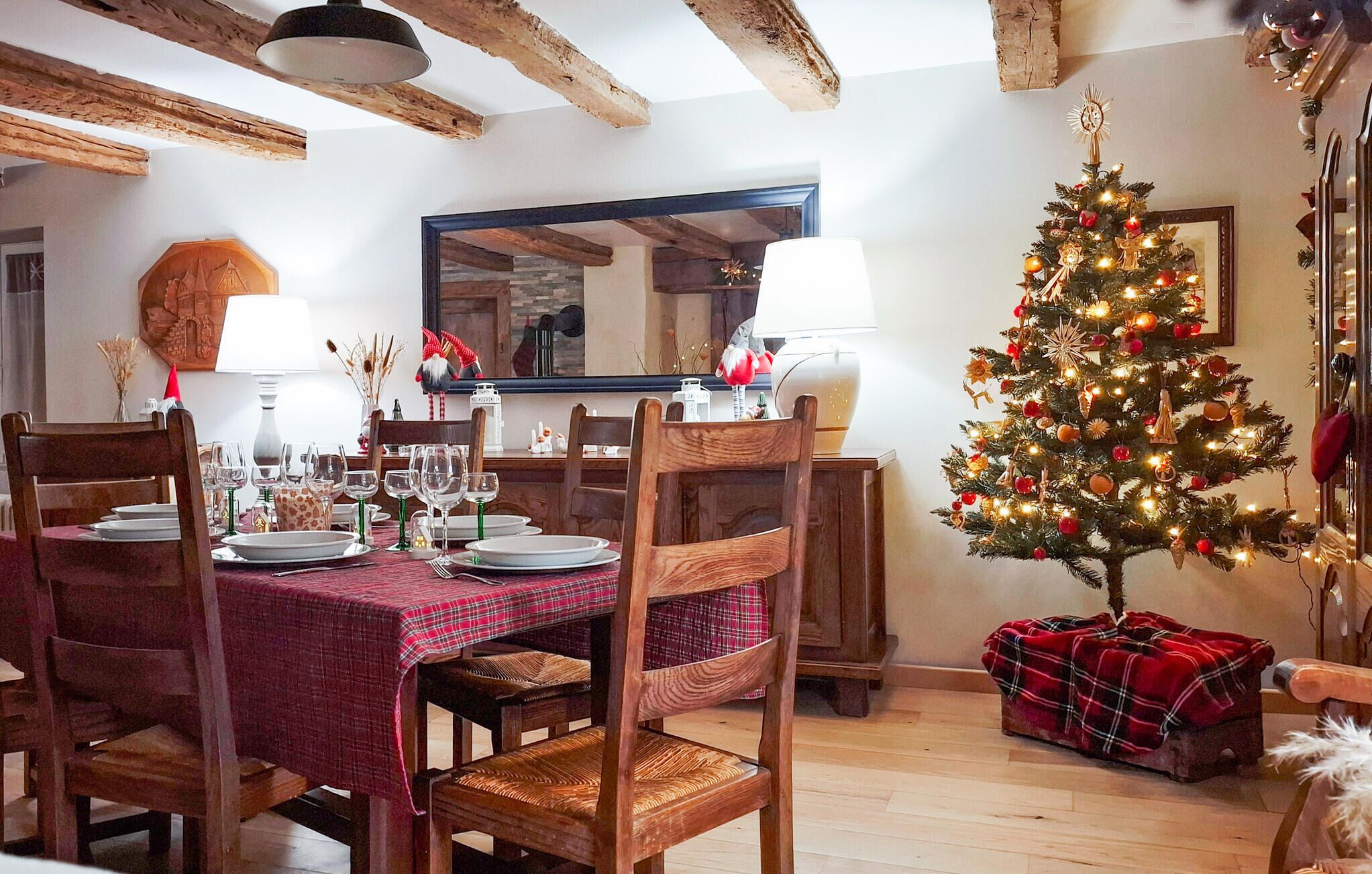 A dining area with a Christmas tree, wooden chairs, a red plaid tablecloth, and a mirror on the wall.