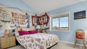 A bedroom with a bed, a nightstand, a chair, a window with blinds, and wall decorations including a 'Welcome Aboard' sign and a lighthouse.